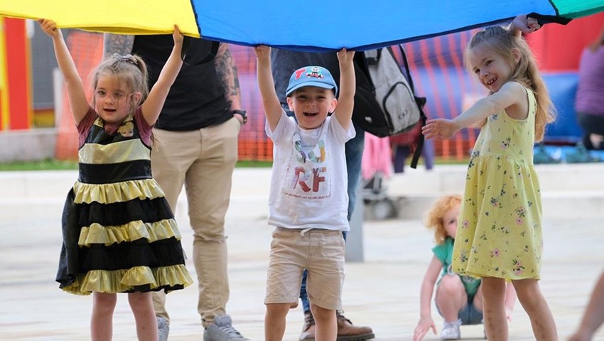 Three children playing with a giant piece of rainbow fabric, smiling.